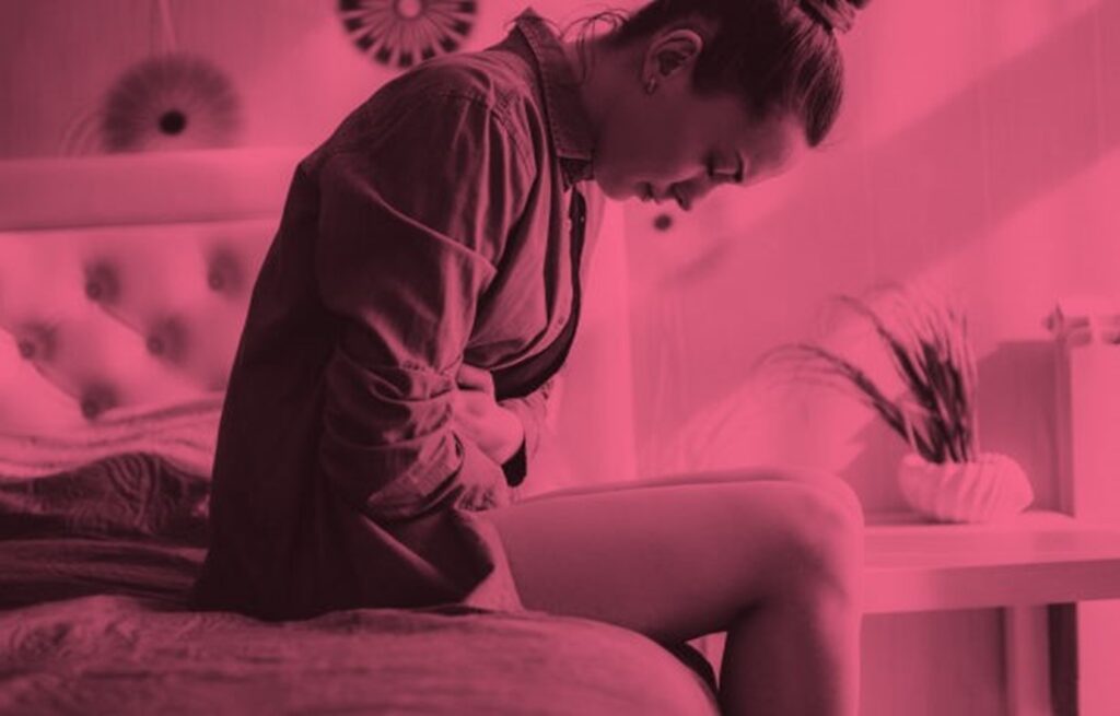 A woman sits on a bed, bent forward, holding her stomach, with a pained expression. The image has a pink filter.