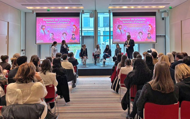 Le public d'une conférence assiste à une table ronde sur scène, sous deux écrans portant l'intitulé « Femmes de sciences, femmes d’impact ».
