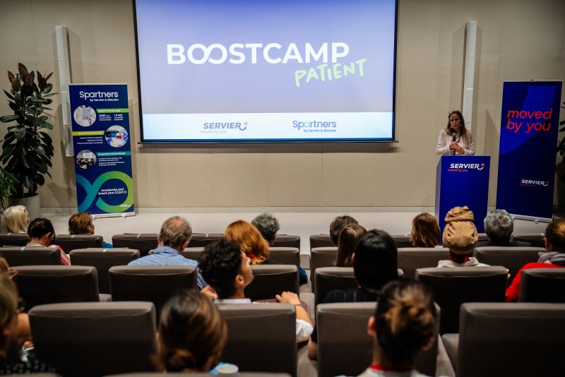 Une femme prend la parole depuis un podium, devant un écran sur lequel s'affiche « Boostcamp Patient », lors d'une présentation devant un public assis.